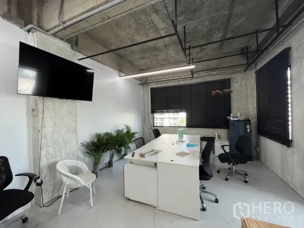 Office Space for rent in Khlong Toei, Bangkok - Loft-style office interior with exposed concrete ceiling, white desks, large windows with black blinds and a wall-mounted TV.