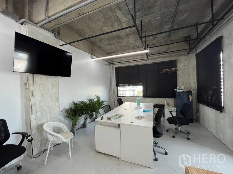 Office Space for rent in Khlong Toei, Bangkok - Loft-style office interior with exposed concrete ceiling, white desks, large windows with black blinds and a wall-mounted TV.