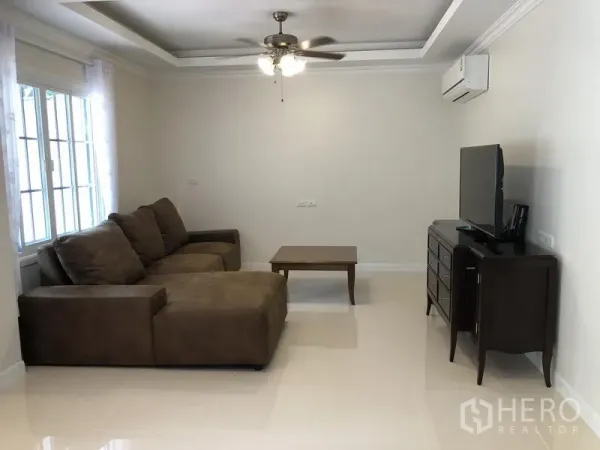 Detached House for rent in Mueang Samut Prakan, Samut Prakan - Cozy living room with brown sectional sofa, TV console and ceiling fan by large windows.