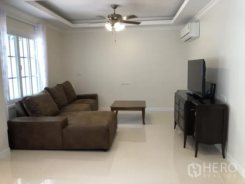 Detached House for rent in Mueang Samut Prakan, Samut Prakan - Cozy living room with brown sectional sofa, TV console and ceiling fan by large windows.