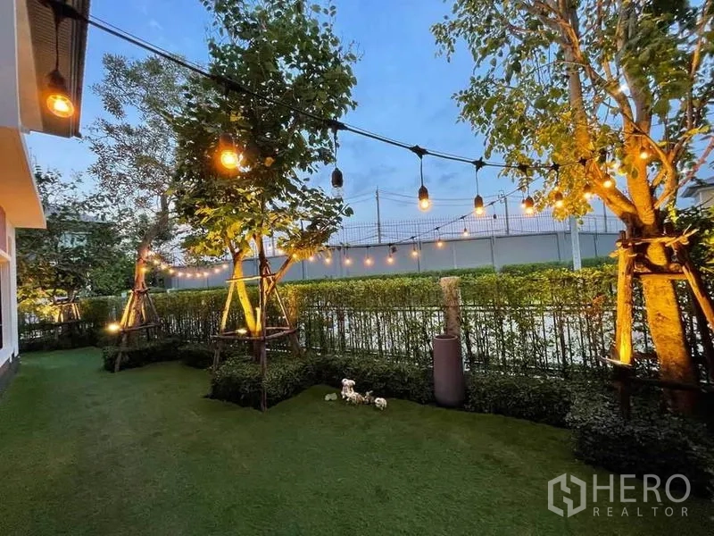 บ้าน ขายในPak Kret, Nonthaburi - สนามหญ้าหลังบ้านติดไฟสตริงไลต์ ต้นไม้ และแนวพุ่มไม้ช่วงเย็น