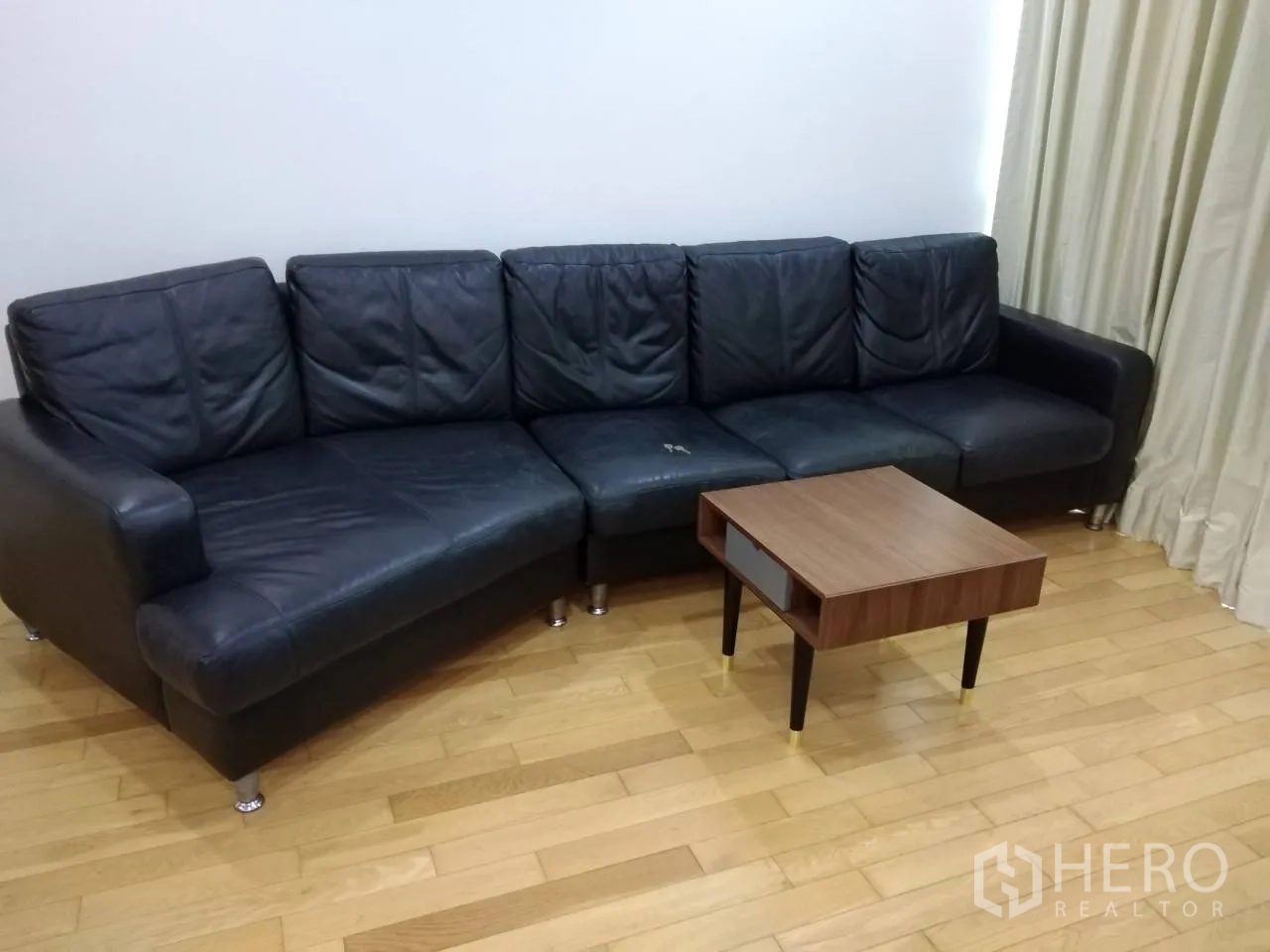 Condominium for rent in Khlong Toei, Bangkok - Black leather sectional sofa with a compact wooden coffee table on polished hardwood flooring.