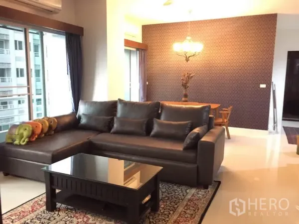 公寓 出租在Bang Kapi, Bangkok - 明亮客厅，深色皮沙发组合、玻璃茶几、纹理特色墙与通往阳台的推拉门。