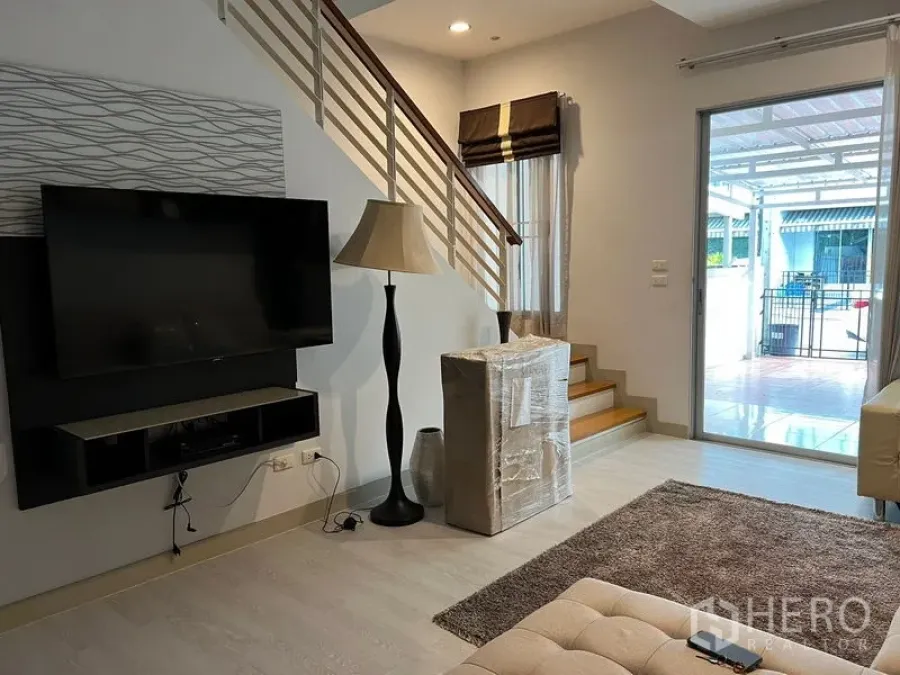 Townhouse for rent in Saphan Sung, Bangkok - Bright living room with wall-mounted TV, floor lamp, staircase and sliding door to a covered patio.