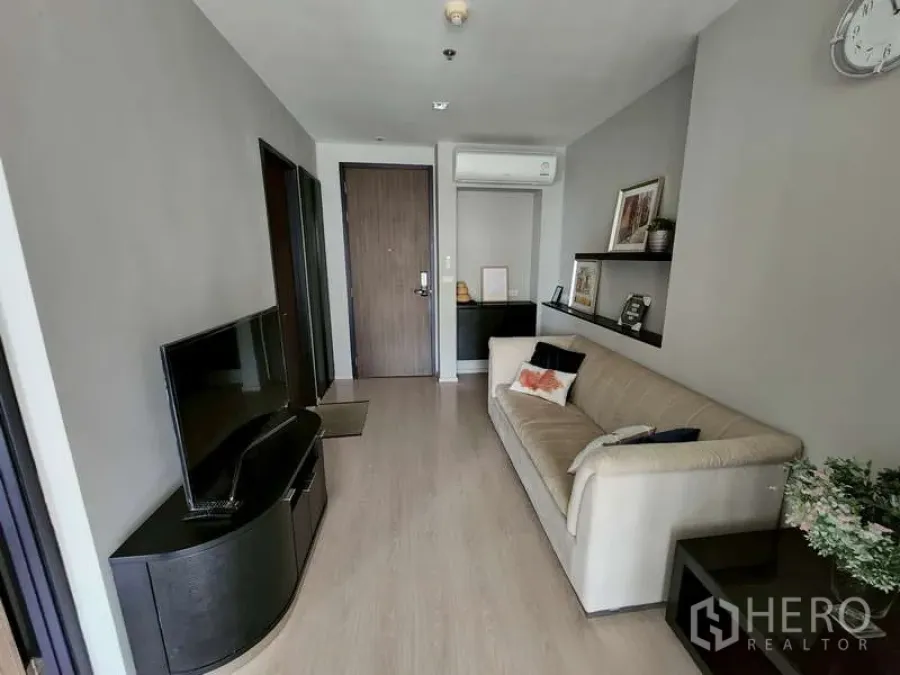 Condominium for sale - Bright living room with TV console, beige sofa, wood flooring, shelves and air conditioning by the entry door.