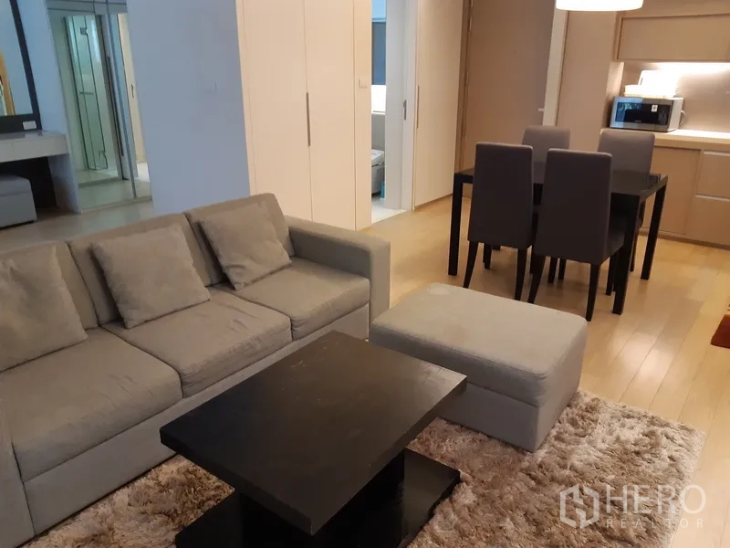 公寓 出租在Watthana, Bangkok - 开放式客厅，灰色沙发与脚凳、黑色茶几与长绒地毯，旁接四人餐桌与一体化厨房。