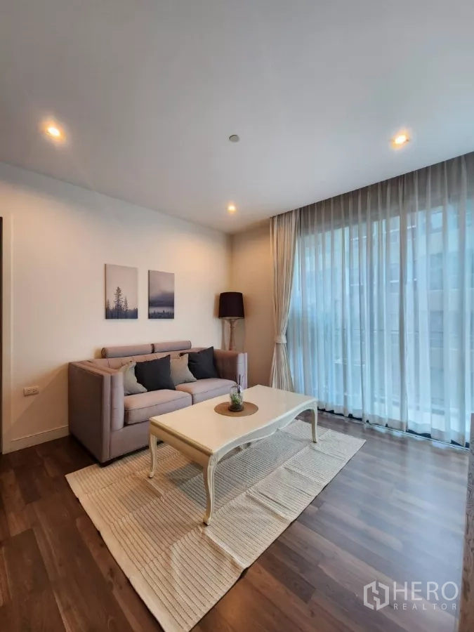 Condominium for rent in Phra Khanong, Bangkok - Spacious living area with sofa, cream coffee table, rug, and wall-to-wall windows.
