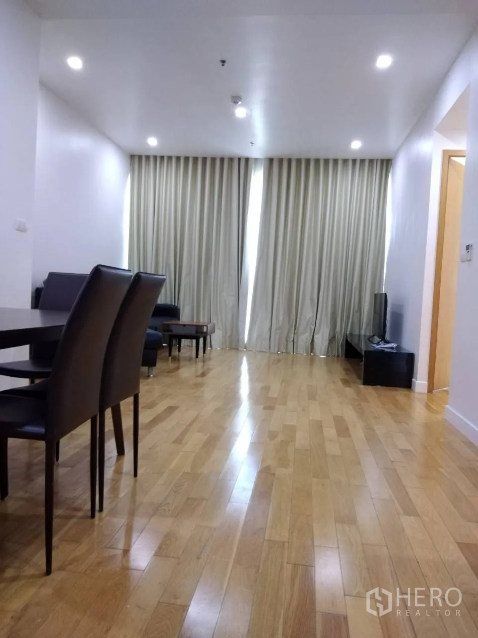 Condominium for rent in Khlong Toei, Bangkok - Spacious living area with wood flooring, ceiling spotlights, and floor-to-ceiling curtains leading to the TV corner.