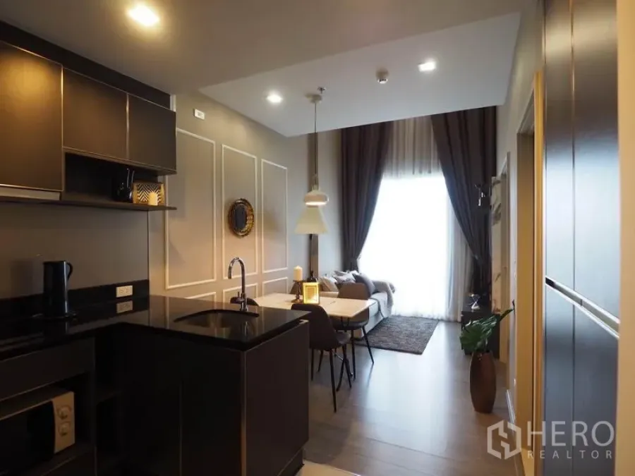 Condominium for rent in Khlong San, Bangkok - Open-plan kitchen with black stone countertop next to dining and living area with tall curtains.