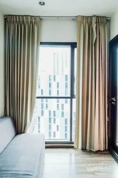 Condominium for rent in Khlong Toei, Bangkok - Living area corner with floor-to-ceiling window, beige curtains and a balcony door.