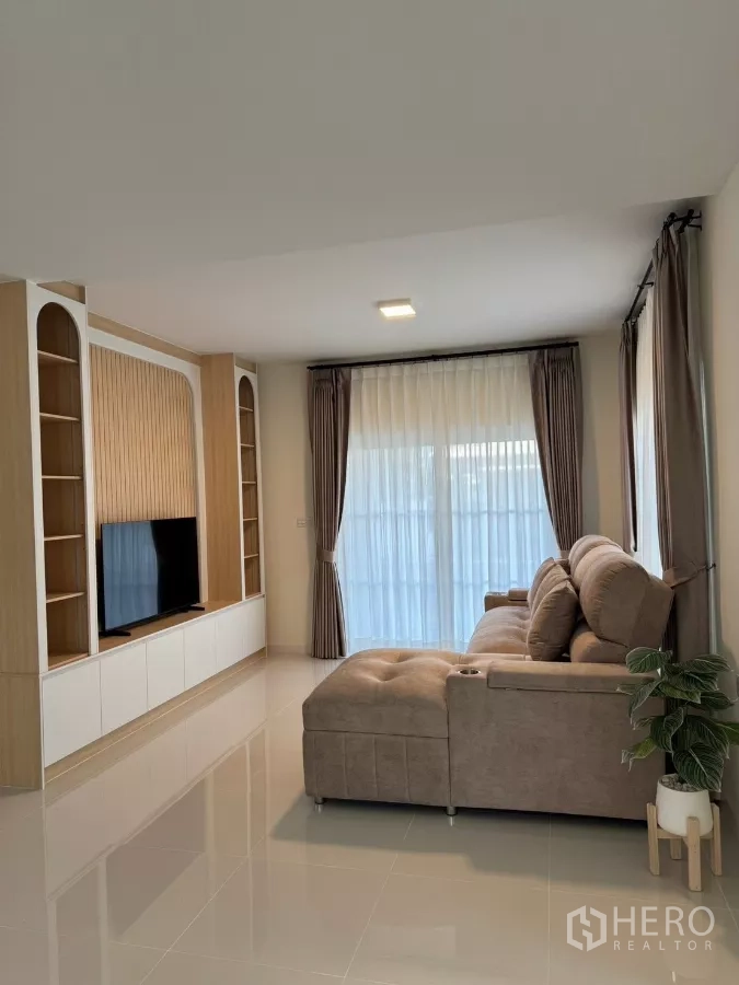 独立式住宅 出租在Bang Phli, Samut Prakan - 客厅配L型沙发与电视背景柜，落地窗帘与亮面地砖。