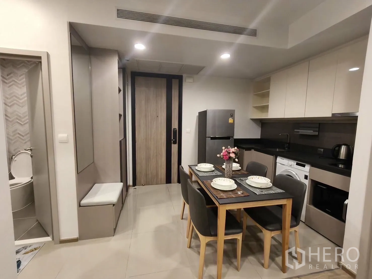 Condominium for rent in Khlong Toei, Bangkok - Entry hall and modern kitchen with a four-seat dining table, refrigerator, and a glimpse of a bathroom with chevron tiles.