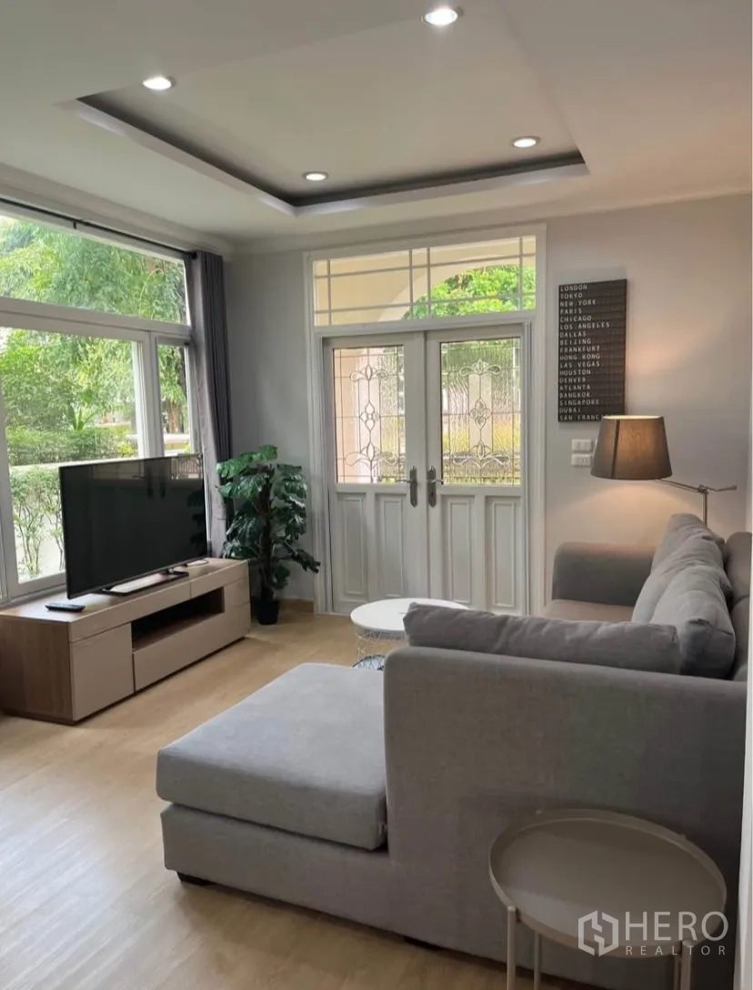 Detached House for rent or sale in Lat Phrao, Bangkok - Bright living room with L-shaped sofa, TV console, large windows and glass double doors.