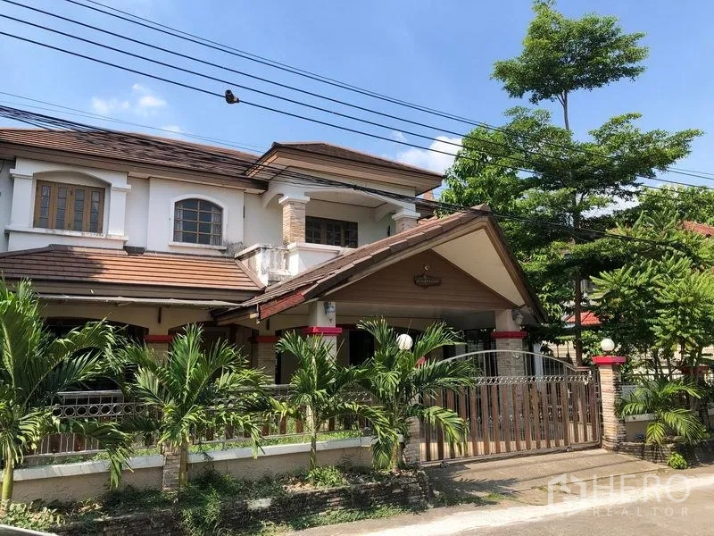 Detached House for sale in Pak Kret, Nonthaburi - Two-storey detached house in Pak Kret, Nonthaburi with tiled roof, balcony, gated fence and a palm-lined front garden under blue sky.