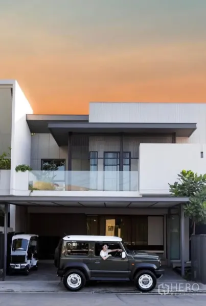 Townhouse for rent or sale in Bang Phli, Samut Prakan - Modern two-storey townhouse facade with glass balcony and covered multi-car parking at sunset.