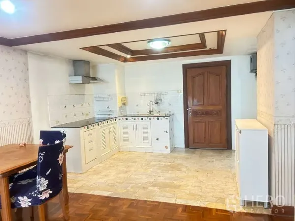 Condominium for sale in Huai Khwang, Bangkok - Open-plan kitchen and dining area with white cabinets, tiled backsplash, range hood, and a solid wooden entry door.