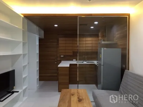 Condominium for rent in Bang Rak, Bangkok - Open-plan living room with built-in white shelving, a glass-partitioned wood kitchen, gray sofa and wooden coffee table.