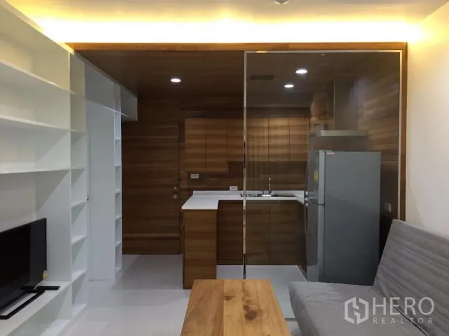 公寓 出租在Bang Rak, Bangkok - 开放式客厅，白色嵌入式置物架，玻璃隔断木质厨房，灰色沙发与木质茶几。