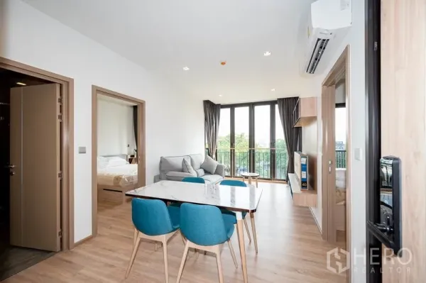 Condominium for rent in Watthana, Bangkok - Bright living and dining area with marble table, blue chairs, sofa and floor‑to‑ceiling windows opening to the balcony.