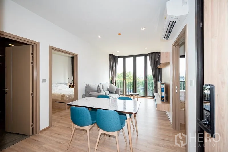 Condominium for rent in Watthana, Bangkok - Bright living and dining area with marble table, blue chairs, sofa and floor‑to‑ceiling windows opening to the balcony.