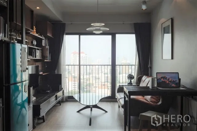 Condominium for rent in Khlong Toei, Bangkok - Living room with large window and skyline view, work desk, sofa and shelving.