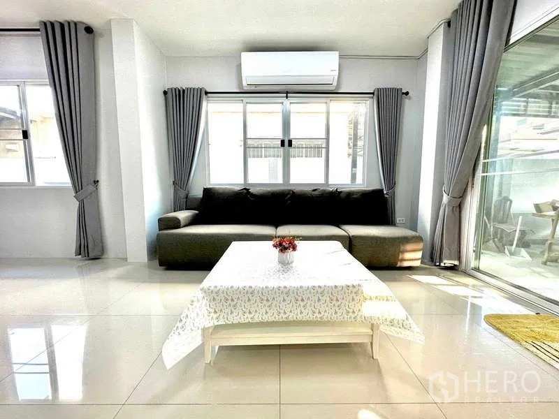 Detached House for rent in Lat Krabang, Bangkok - Air-conditioned living room with L-shaped sofa, large windows and tiled floor.