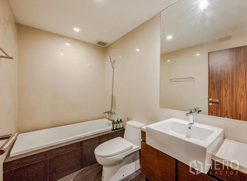 Condominium for sale in Khlong Toei, Bangkok - Bathroom with bathtub, shower, white basin, large mirror, and warm neutral tiles.