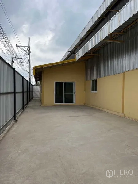 货栈 出租在Lat Krabang, Bangkok - 金属外墙仓库旁的混凝土车道与安防围栏，端部为玻璃推拉门入口。
