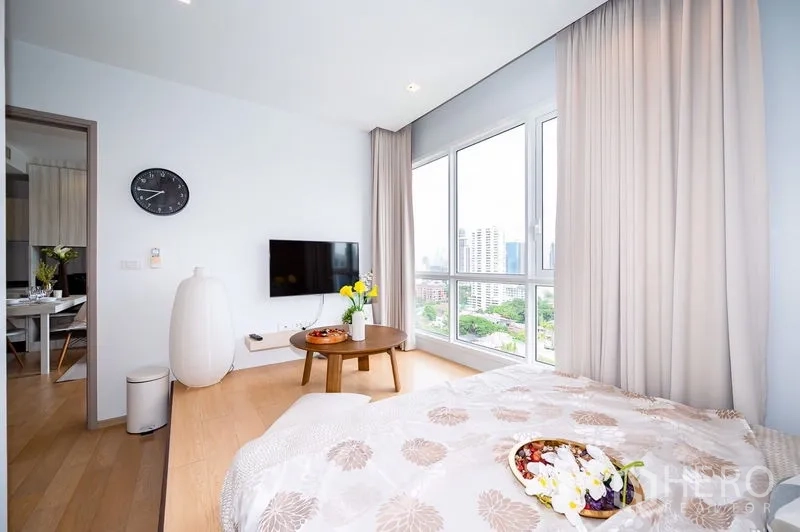 公寓 出租或出售在Watthana, Bangkok - 卧室配全景落地窗、墙挂电视和浅色窗帘，俯瞰绿意与楼群