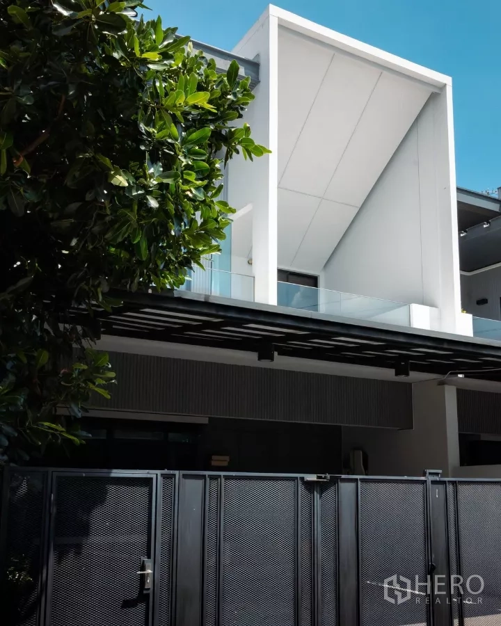 Detached House for rent or sale in Bang Phli, Samut Prakan - Modern detached house facade at VIVE Bangna with white geometric balcony and black front gate under a tree.