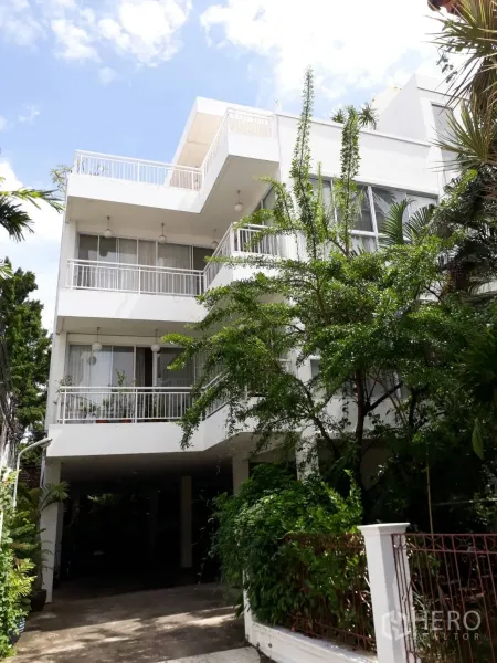 公寓 出租在Watthana, Bangkok - 白色低层公寓外观，带宽阔阳台与大窗户，绿植环绕并设有架空车位。