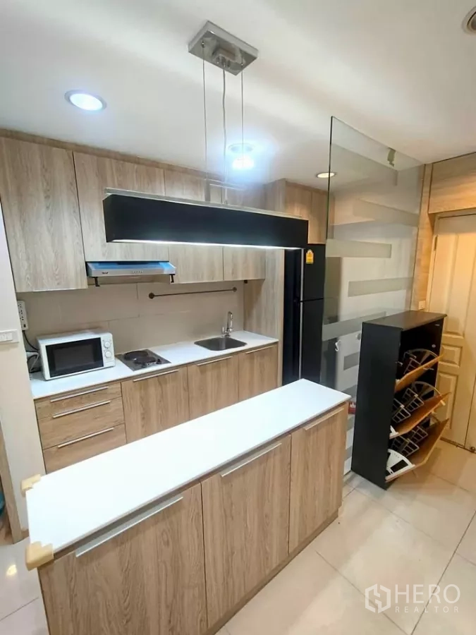 Condominium for rent in Khlong Toei, Bangkok - Fitted kitchen with wood cabinetry, long island counter, range hood, microwave, refrigerator, and a glass partition.