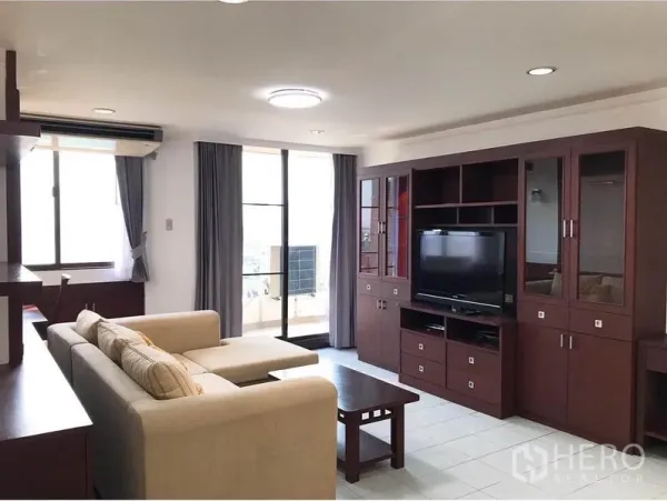 Condominium for rent or sale in Watthana, Bangkok - Bright living room with L-shaped sofa, dark wood TV wall and sliding doors to balcony.