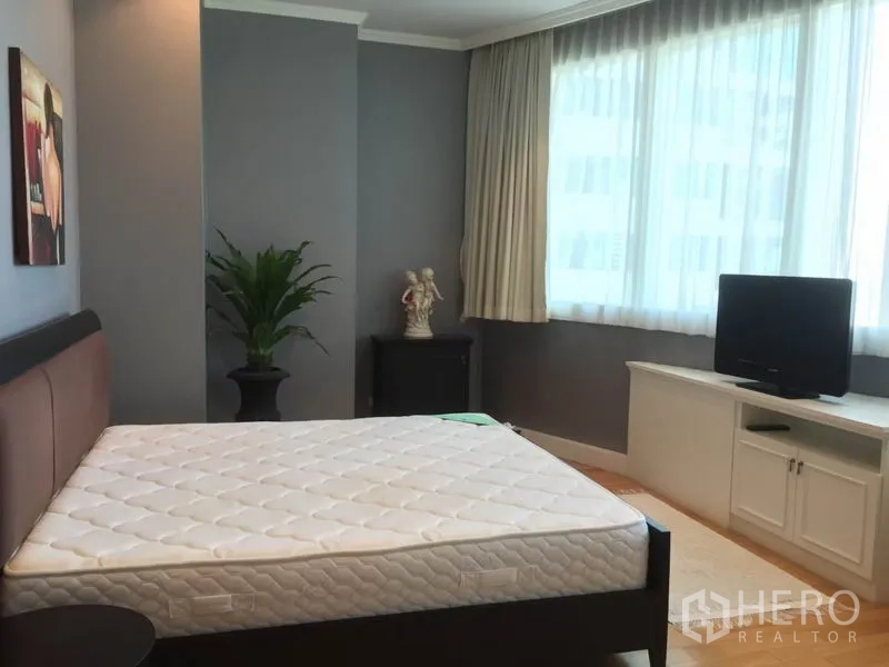 Condominium for rent in Khlong Toei, Bangkok - Primary bedroom with large window, gray accent wall, bed frame, TV on built‑in cabinet and potted plant.