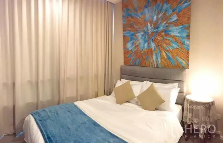 คอนโด ให้เช่าหรือขายในWatthana, Bangkok - ห้องนอนโทนอุ่น มีผ้าม่านยาว โคมไฟข้างเตียง และภาพศิลปะสีสันเหนือเตียง