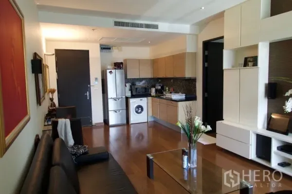 Condominium for sale in Pathum Wan, Bangkok - Open-plan living room with wooden floors, built-in shelving, and a kitchenette with fridge and washing machine.