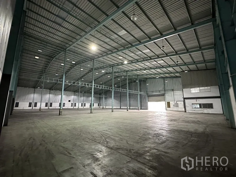Warehouse for rent in Mueang Samut Prakan, Samut Prakan - Open warehouse floor with high clearance, skylights, roll-up door and a small built-in office section.