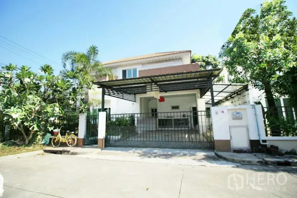 Detached House for rent or sale in Lat Krabang, Bangkok - Front view of a two-storey detached house with gated covered carport and mature trees in Lat Krabang.
