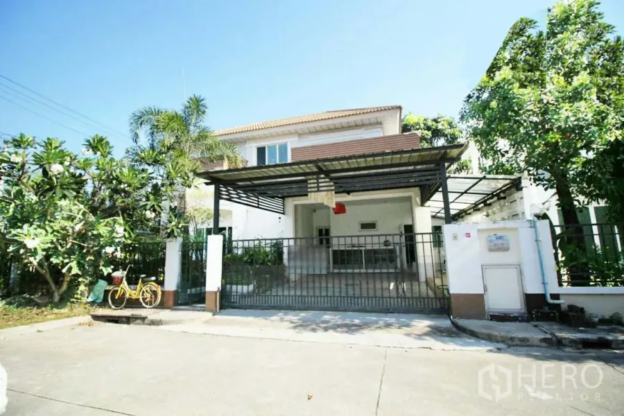 Detached House for rent or sale in Lat Krabang, Bangkok - Front view of a two-storey detached house with gated covered carport and mature trees in Lat Krabang.