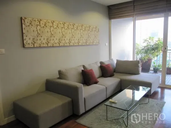 Condominium for rent in Khlong Toei, Bangkok - Cozy lounge featuring a gray sectional, red accent cushions, and a balcony with potted greenery.