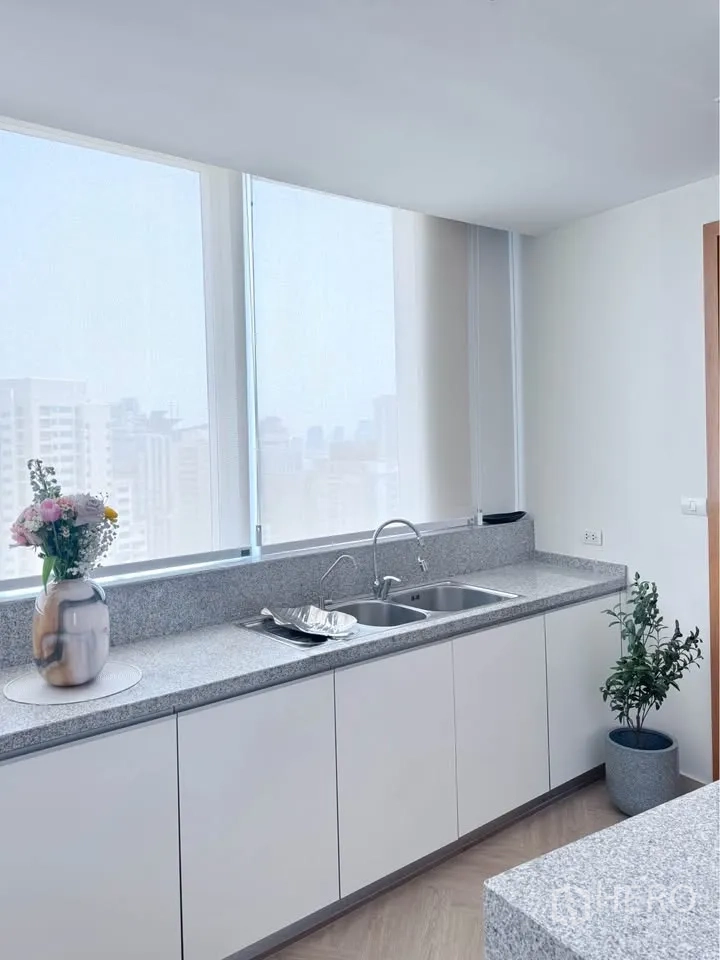 Condominium for rent or sale in Khlong Toei, Bangkok - Bright kitchen with long granite counter, double sink, and large windows showing city views.