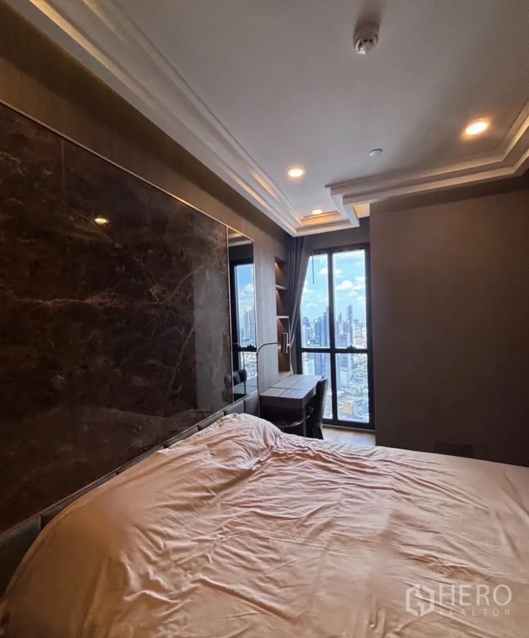 Condominium for rent in Bang Rak, Bangkok - Bedroom with a marble feature wall, built-in desk, and floor-to-ceiling window overlooking the city skyline.