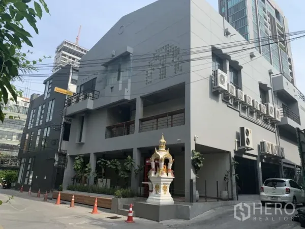 Retail Space for rent in Khlong Toei, Bangkok - Modern gray low-rise commercial building exterior with balconies, air-conditioning units, a spirit house and parking area.