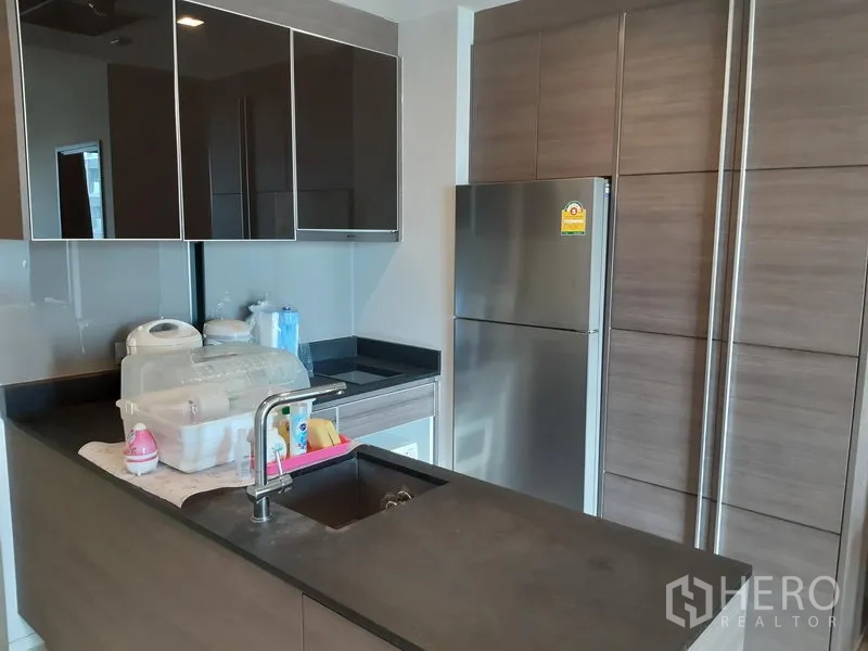 Condominium for rent in Khlong Toei, Bangkok - Modern kitchenette with dark countertops, glossy cabinets and a stainless-steel refrigerator.