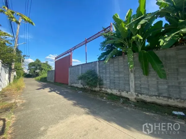 Warehouse for rent in Suan Luang, Bangkok - Quiet lane with high perimeter wall and large red sliding gate leading to the warehouse.