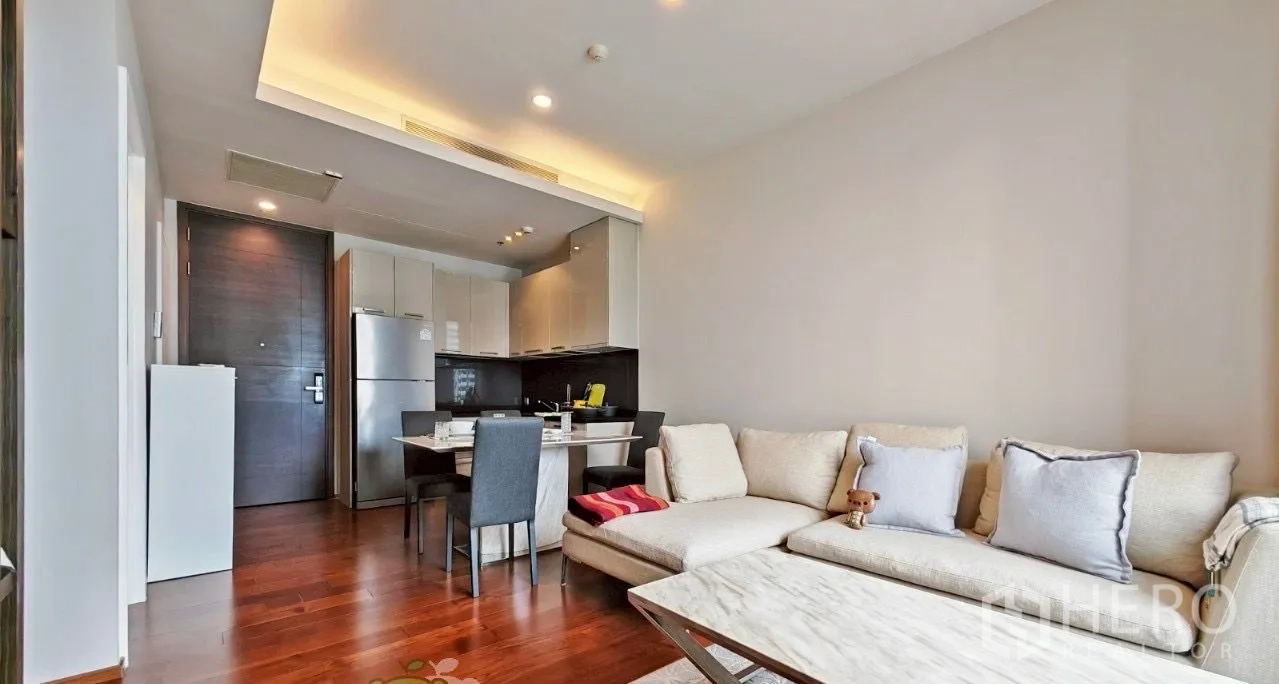 公寓 出租在Watthana, Bangkok - 开放式起居区，沙发、餐桌与紧凑厨房一体，暖色吊顶灯和木地板。