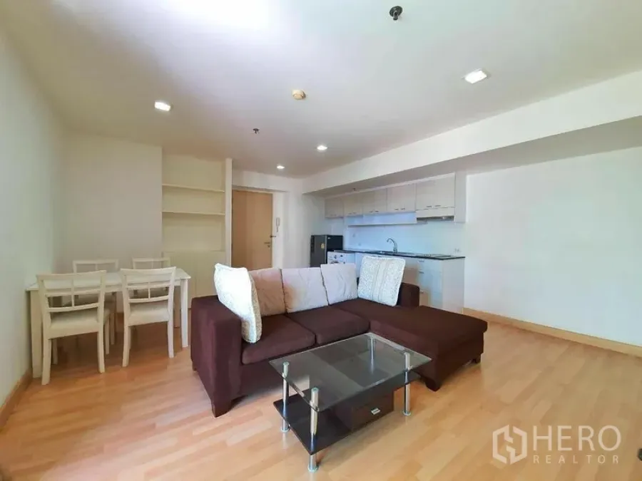Condominium for rent in Din Daeng, Bangkok - Living room view showing L-shaped sofa, glass coffee table, built-in shelving, and long kitchen counter.