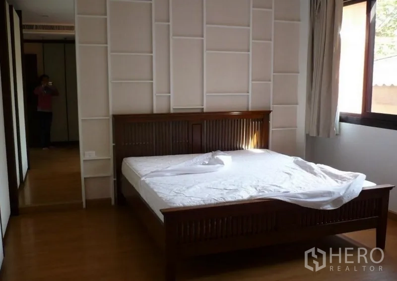 Detached House for rent in Khlong Toei, Bangkok - Bright bedroom with wooden bedframe, feature wall panels and a window bringing in natural light.