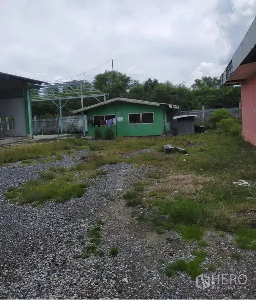 Factory for rent or sale in Samut Prakan - Open gravel yard with a small green office building and surrounding factory structures for parking and loading.