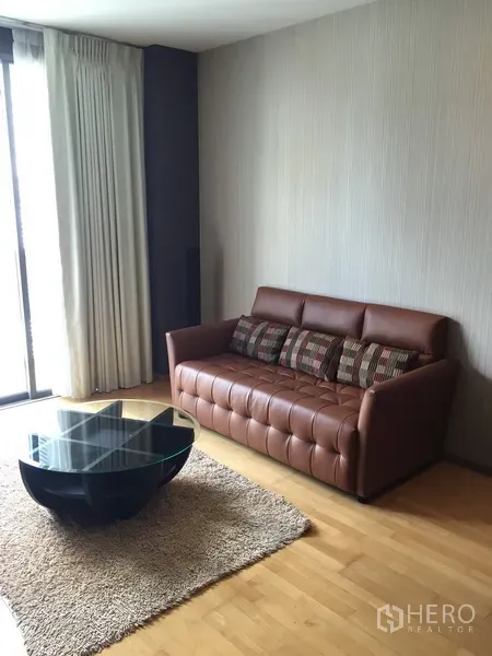 Condominium for rent or sale in Khlong Toei, Bangkok - Cozy lounge with brown leather sofa, glass coffee table and floor-length curtains.